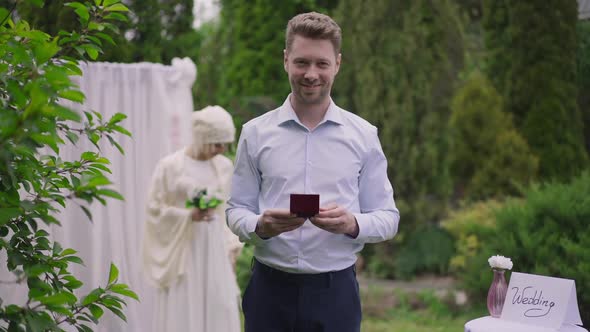 Portrait of Happy Caucasian Groom Holding Rings Looking at Camera Smiling As Blurred Gorgeous Middle