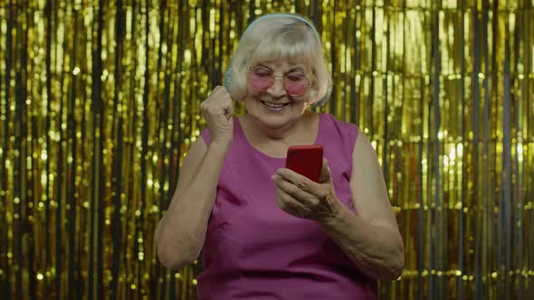Stylish Happy Senior Old Woman Holding Cell Phone, Listening To Music in Headphones and Dancing