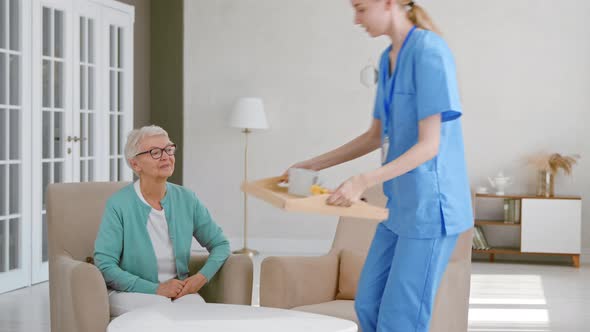 Positive young nurse carries tray with breakfast to senior woman