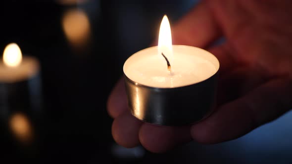 A hand holding a small tealight candle with the flames of many burning in the background during a ca