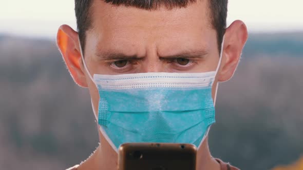 Man in Protective Face Mask Looks at Smartphone Holding It in Hands on Street