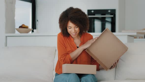 Excited African Woman with Curly Hair Customer Female Buyer Opening Parcel Box at Home Sitting on