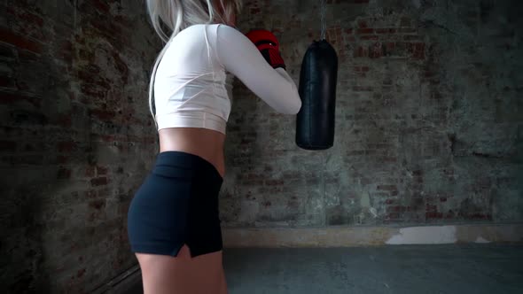 Blonde Slender Female Boxer in Shorts and a Top, She Is in Gloves Punching a Punching Bag, Training