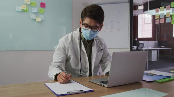 Asian male doctor wearing mask sitting at desk in meeting room making notes