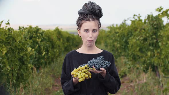 Young Woman with Dreadlocks Biting Black Grapes From Her Hands Standing at Vineyard