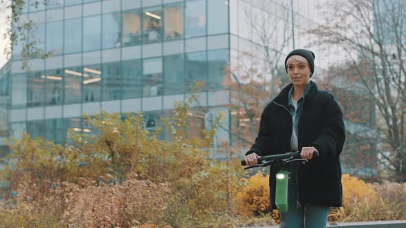 Young Woman in Winter Black Jacket with Hat Riding Electic Scooter in the City Park. Ecological