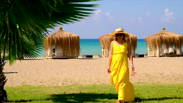 A Woman in a Hat and Dress Walks on the Beach