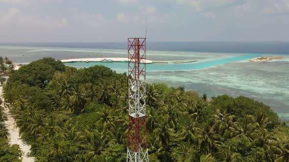 A high cell tower stands in a town on the ocean surrounded by green trees against a beautiful sky