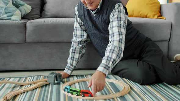 Retired Old Man Playing with Wooden Toy Cars Sitting on Floor in Apartment