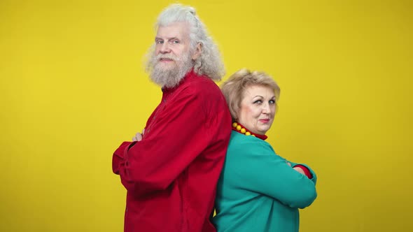 Confident Senior Man and Woman Standing Back To Back on Yellow Background Turning To Camera and