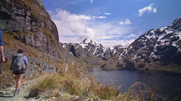 Static, hikers walk above alpine lake through snow capped mountain landscape, Routeburn Track New Ze