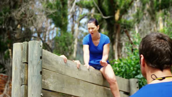 Female trainer assisting fit woman to climb over wooden wall during obstacle course