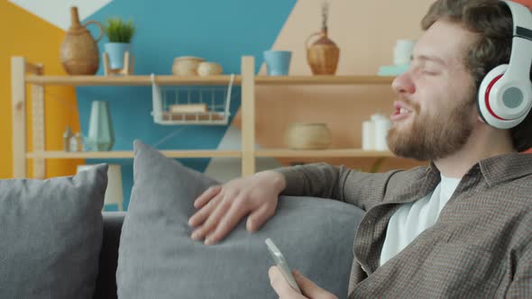 Happy Male Student Enjoying Music Through Headphones Holding Smartphone Indoors in Apartment