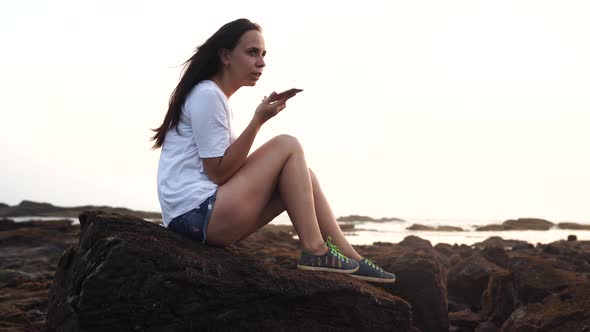 A beautiful young woman is sitting on the rocks and recording audio messages