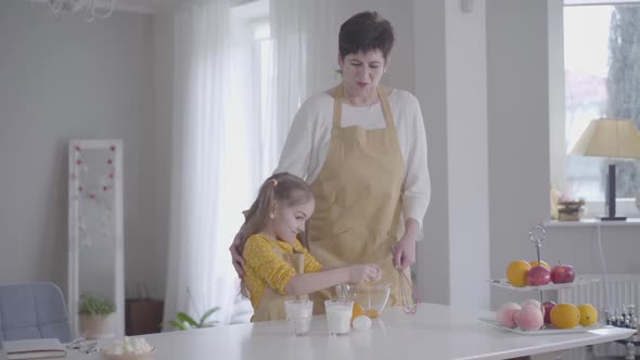 Little Cute Caucasian Girl Breaking Egg Into Bowl and Smiling. Happy Granddaughter Helping