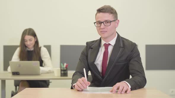 Confident Young Male Manager in the Suit Sitting at the Desk in the Foreground Looking in the Camera