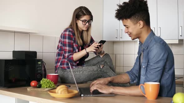 Young Happy Couple of Hipsters Sitting in the Kitchen Both Using Some Devices Like Laptop Smartphone
