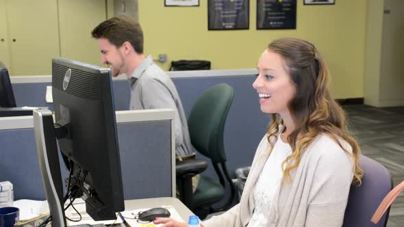 Two young professionals talking and laughing while working on computers in cubicles