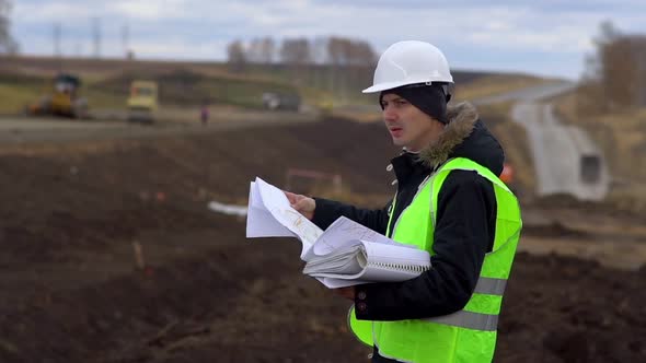 The Worker Looks at the Drawings on the Background of the Road Under Construction