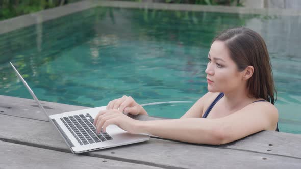 A young mixed-race girl in a blue swimsuit is working at a laptop while standing in the pool.