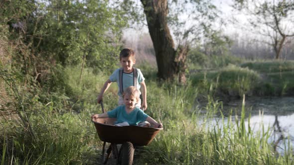 Children Rest in the Countryside, Cheerful Cute Boy Fooling Around with His Brother in Wheelbarrow