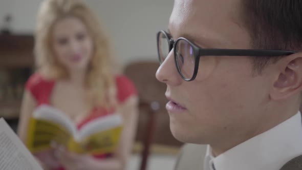 Young Geeky Man in Glasses Reading the Book and Falling Asleep in Foreground While Curly Blond Woman