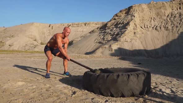 Muscle Athlete Strongman Man Hits a Hammer on a Huge Wheel in the Sandy Mountains