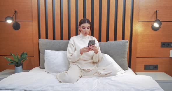 Young Woman Sits on the Bed Female Playing Game on a Smartphone and Wins the Game Emotions of