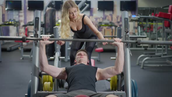 Fit Slim Blond Personal Trainer Helping Sportsman Lifting Barbell in Gym Lying on Exercise Bench