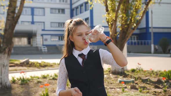 Schoolgirl Teenager in School Uniform Drinks Water From a Plastic Bottle Near the School