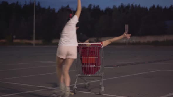 Cheerful Young Woman on Roller Skates Riding Joyful Man Sitting in Shopping Cart. Portrait of Happy