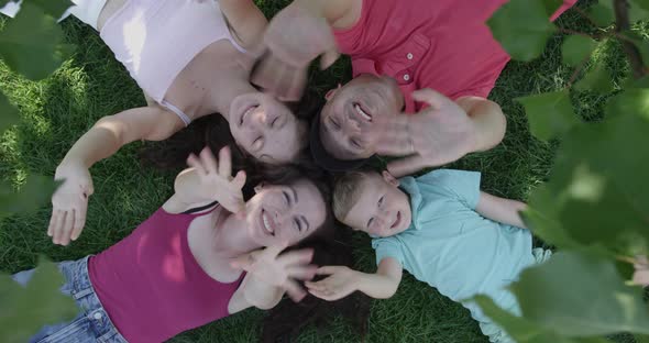 Top View Of Happy Family Waving Hands At Camera And Smiling Laying Down In A Lawn Under A Tree