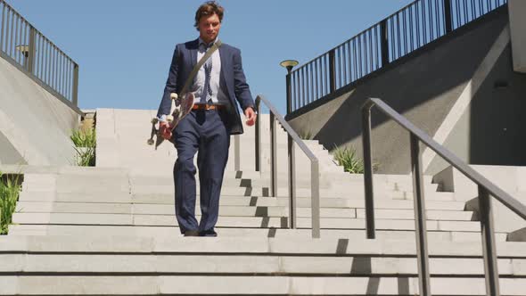 Caucasian businessman walking downstairs, holding skateboard on sunny day