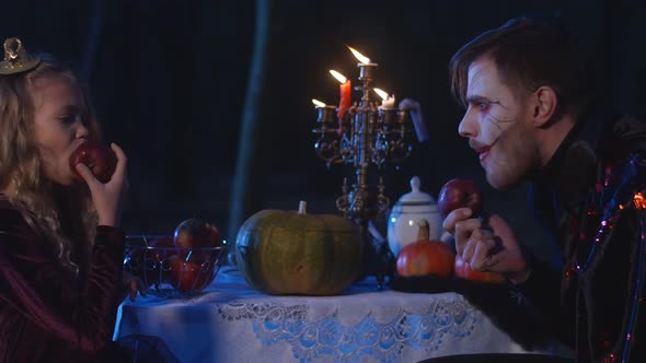 Little Girl and Vampire Sitting in Darkness Eating Apples with Candles Burning at Table at