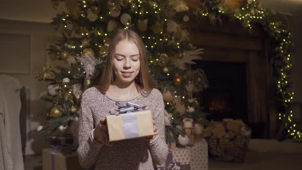 Pretty Smiling Girl Gives the Christmas Present Box To Camera, Standing at Decorative Lighting at