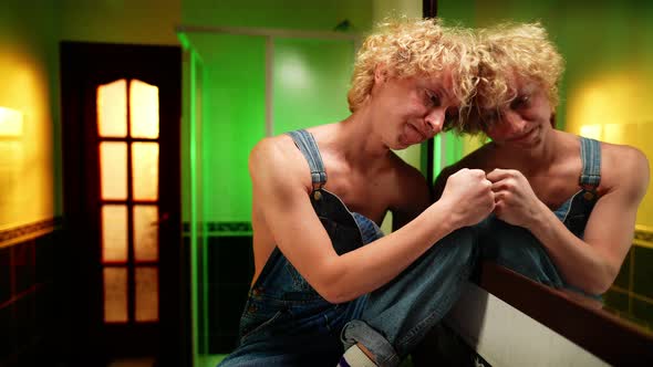 Depressed Queer Man Hitting Mirror with Fist Sitting in Bathroom at Home Indoors