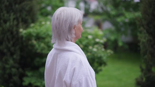 Happy Mature Caucasian Woman in White Bathrobe Standing on Backyard Admiring Nature