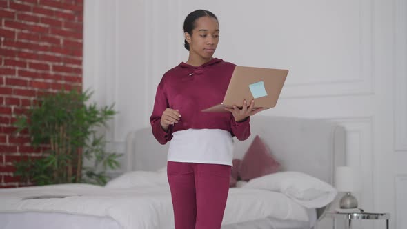 Portrait of Slim Young Woman Standing in Bedroom Holding Laptop in Hand Typing on Keyboard Thinking