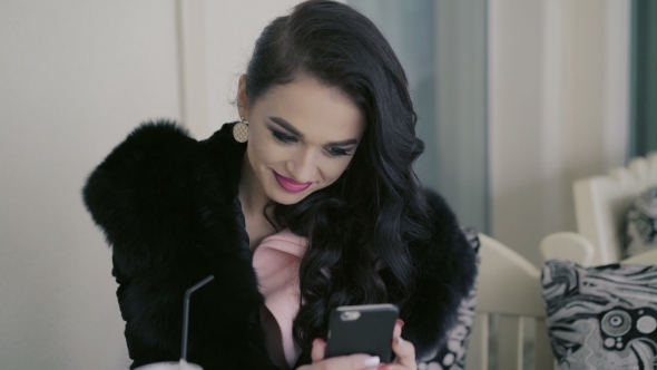 Smart Brunette in Expensive Fur Drinking Juice, Looking on The Phone In Cafe. alt