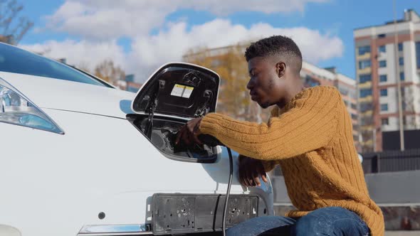 African American Male Driver Connects Electric Car to Power System to Charge Car Battery
