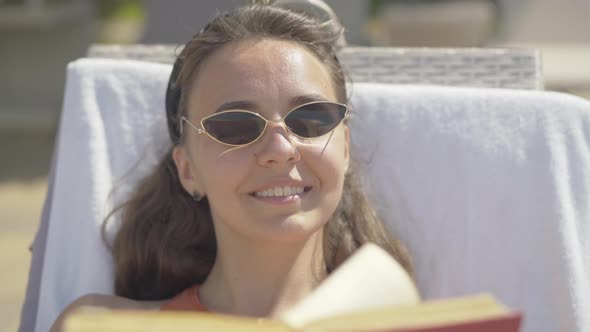 Close-up of Young Positive Woman in Sunglasses Reading Book at Luxurious Resort. Portrait of