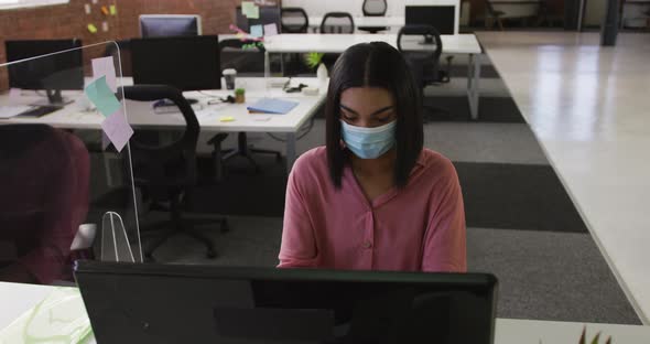 Mixed race businesswoman wearing face mask sitting at desk using computer