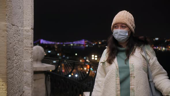 Woman in Mask Leaning at Railing with Istanbul Cityscape Behind at Night