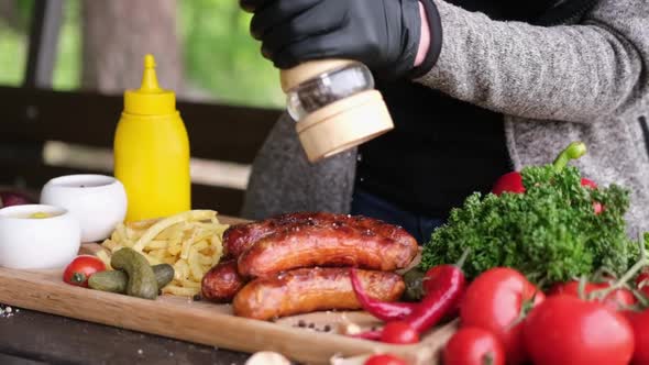 Pour Grinded Pepper on Tasty Grilled Sausages on Wooden Serving Board with French Fries and Sauces