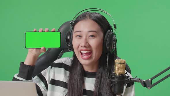 Smiling Asian Woman With Headphone Reviewing Green Screen Mobile Phone While Sitting On Green Screen