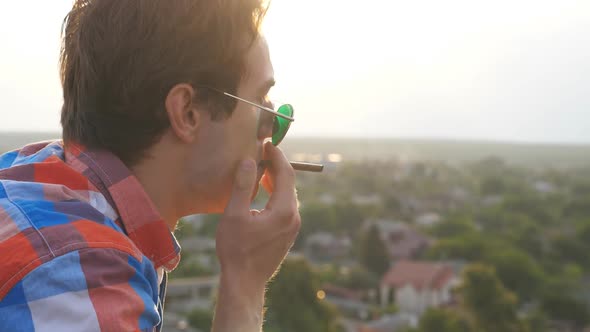 Profile of Handsome Man in Sunglasses Standing on Roof and Smoking Cigarette with Cityscape at