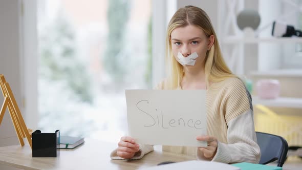 Middle Shot of Sad Beautiful Woman with Taped Mouth Holding Paper with Silence Written Looking at