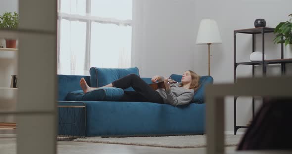 Attractive Young Girl with Blond Hair Is Resting on a Soft Sofa in a Light Living Room