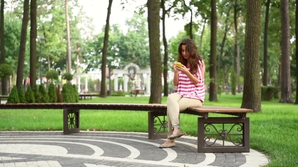 Beautiful Woman Sitting On a Bench In The Park With Smartphone