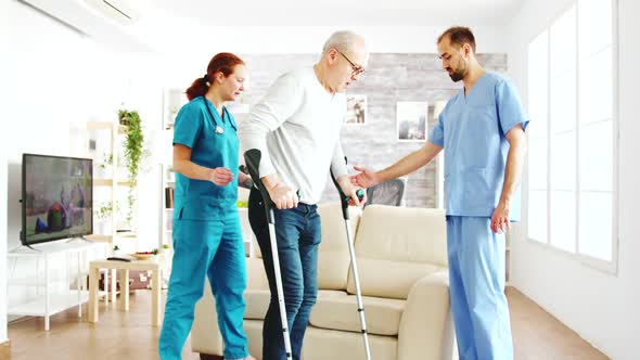 Two Nurses Helping an Old Disabled Man with Crutches To Walk in His Room